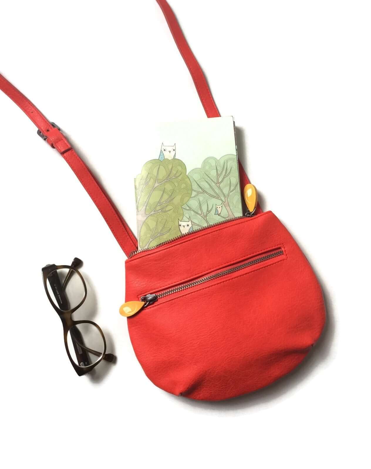 Red vegan leather crossbody bag with adjustable strap, wood zip pullers, bright orange lining, and zippered compartments next to reading glasses.