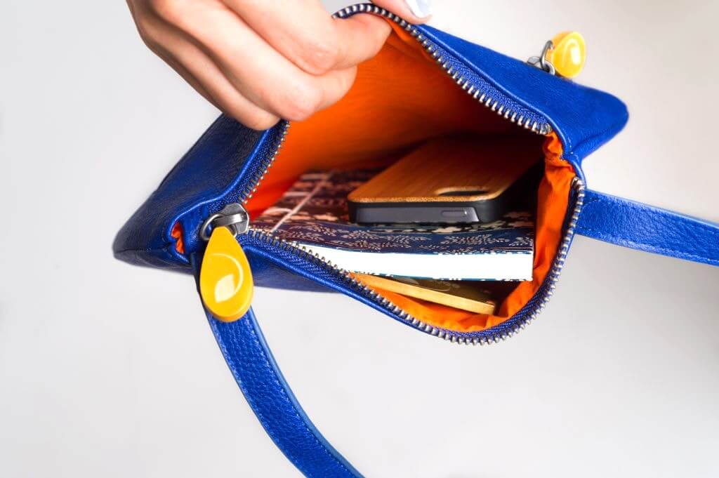 Open blue Willoughby Crossbody Bag showing bright orange lining and essentials like phone, wallet, and book. Vegan leather purse with zippered compartments.