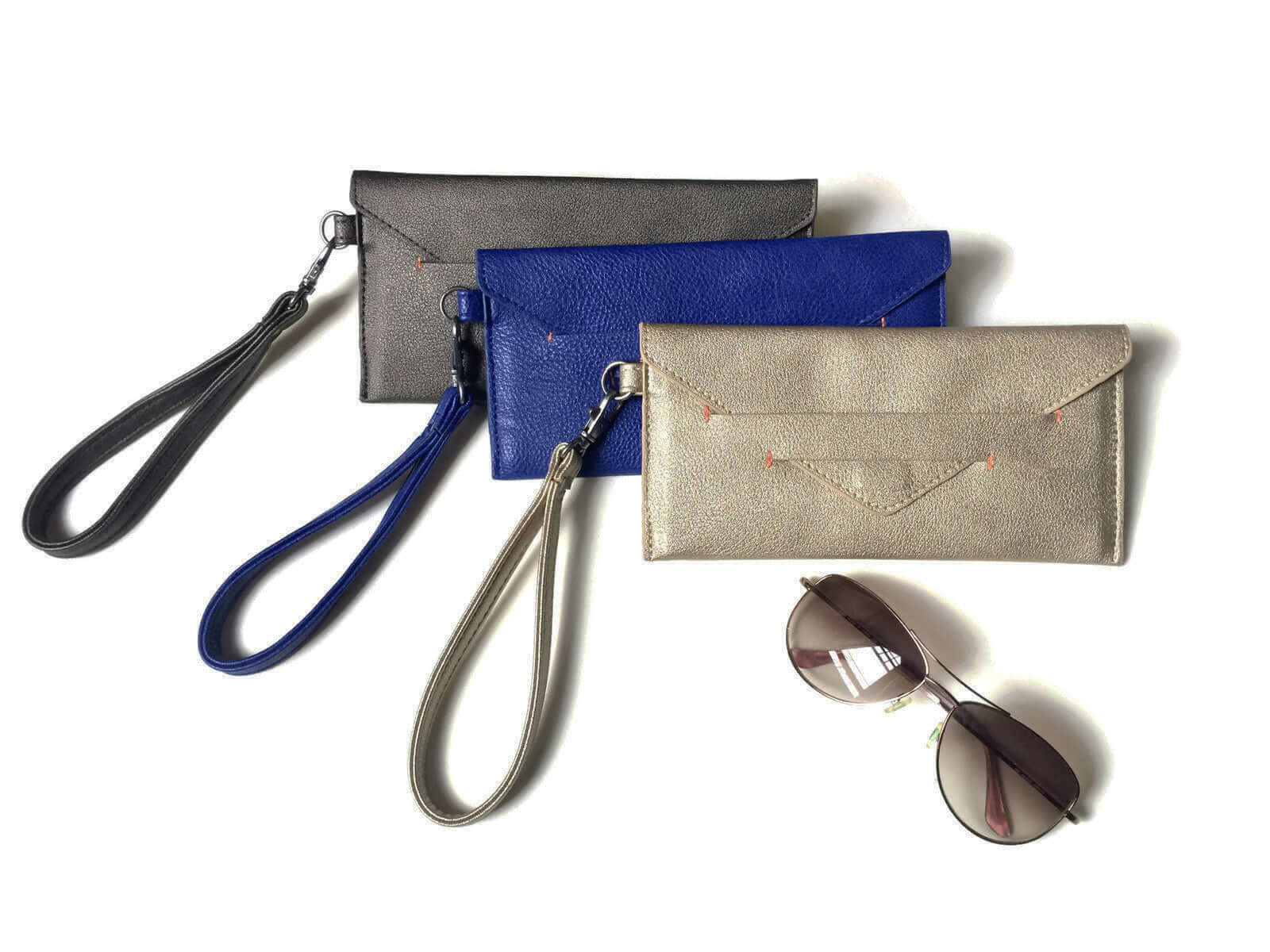 Three Delancey Wristlet Wallets in grey, blue, and beige with detachable wrist straps, lying next to a pair of sunglasses.