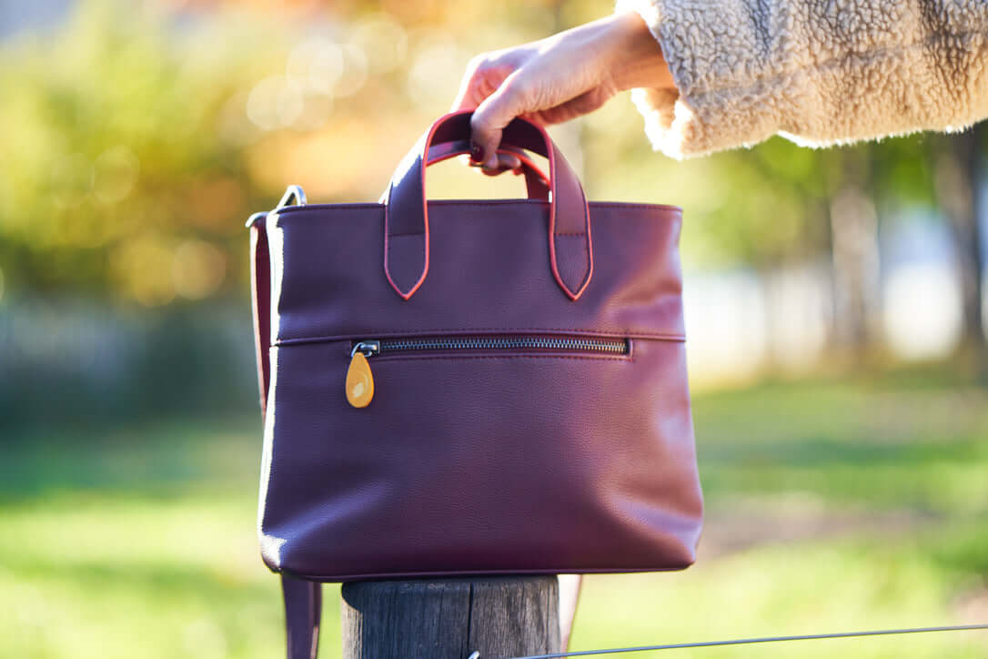 Hand holding Atlantic Crossbody Satchel in vegan leather with orange-edged handles and outside pocket in outdoor setting.