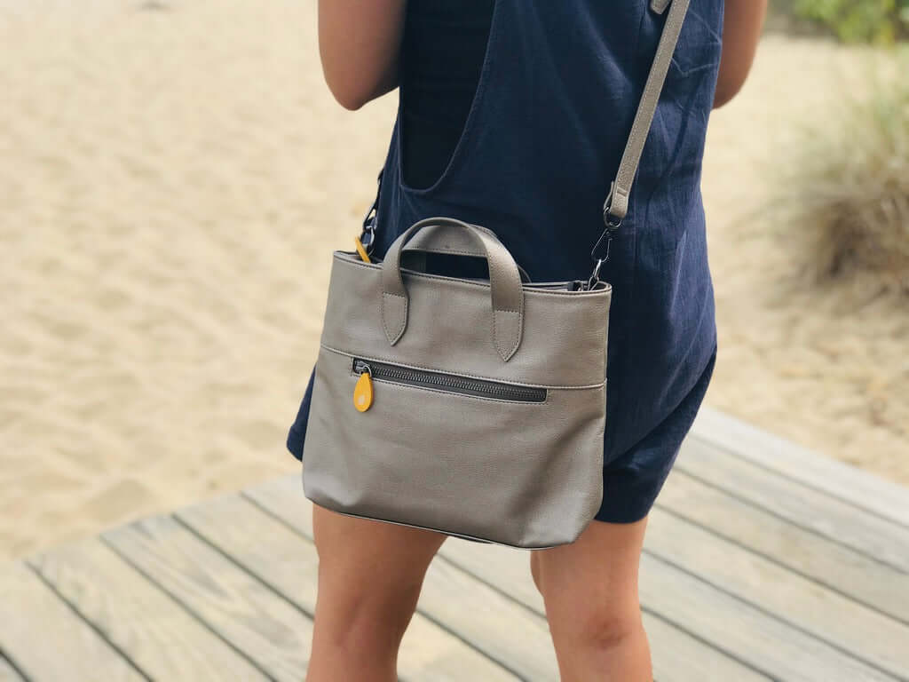 Person carrying gray Atlantic Crossbody Satchel with orange zip puller, vegan leather, and outside pocket, perfect for everyday essentials.