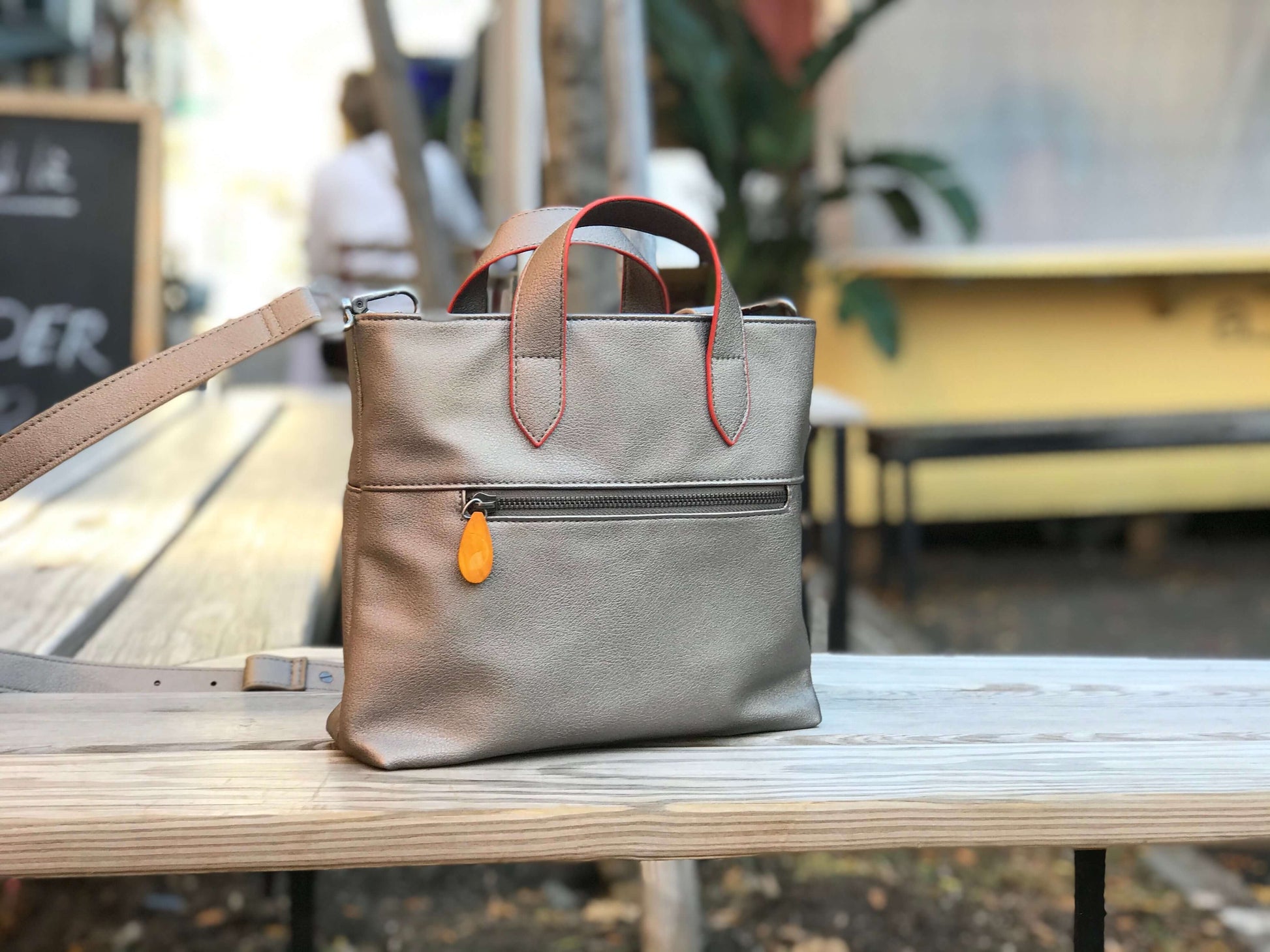 Atlantic Crossbody Satchel in vegan leather with wood zip puller, orange-edged handles, and outside pocket displayed on a wooden bench.