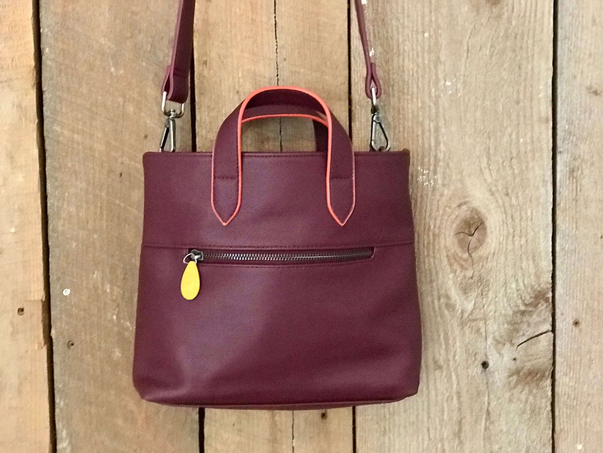 Atlantic Crossbody Satchel in burgundy vegan leather with orange accents shown hanging against a wooden background.