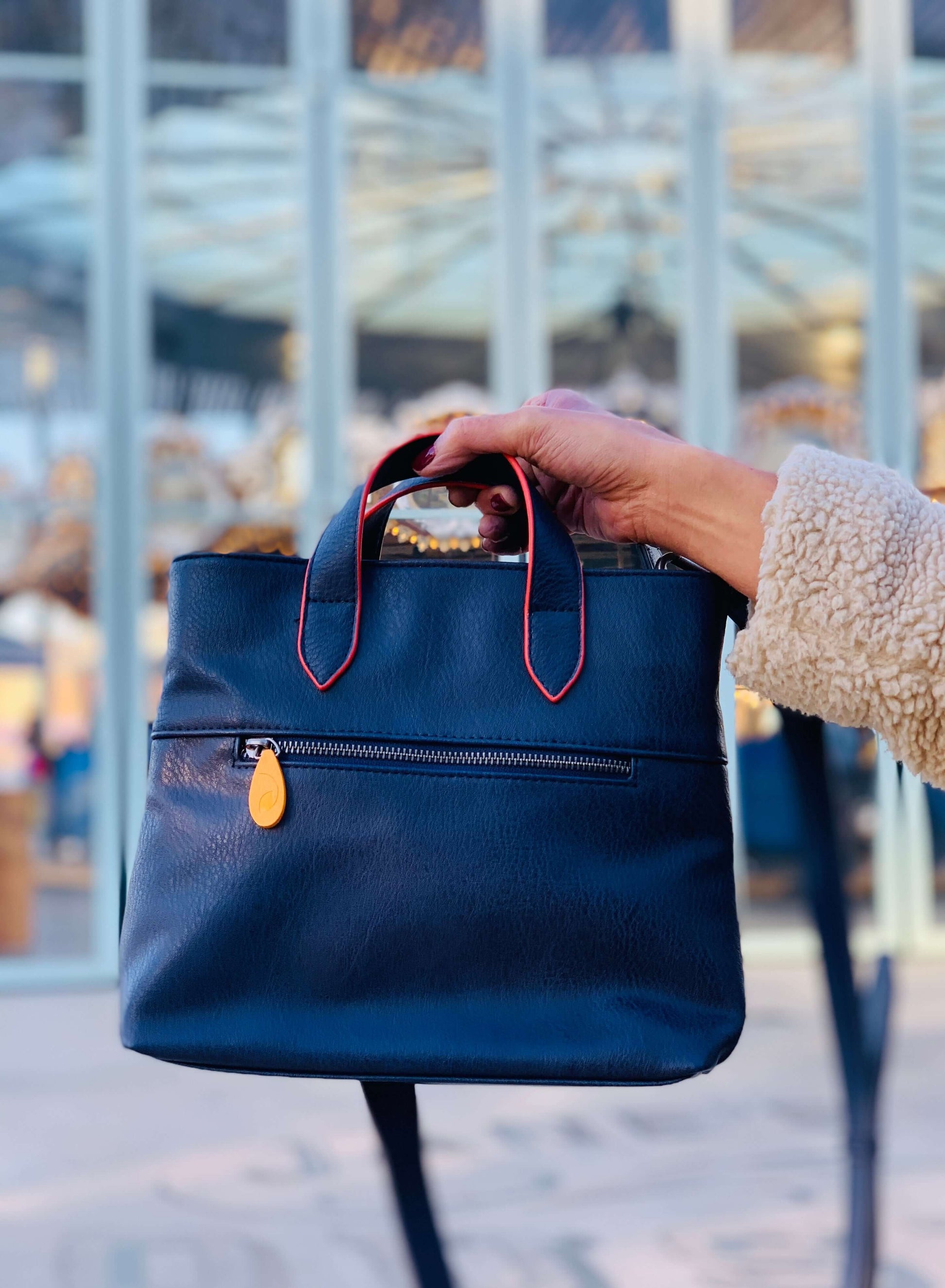 Stylish black Atlantic Crossbody Satchel with orange details and wood zip puller, held against a modern glass building background.